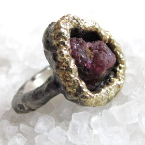 Puede incluir: Anillo artesanal con una gema roja oscura en bruto engastada en un bisel texturizado dorado. La banda del anillo es de color plateado contrastante, creando un diseño único y orgánico. La gema mide aproximadamente 1,3 cm de diámetro.