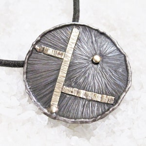 May include: A silver pendant necklace with a round, textured disc featuring a gold geometric design. The disc has a textured surface and a small gold dot in the center.
