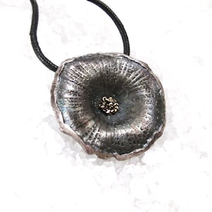 May include: A silver pendant necklace with a textured, flower-shaped design and a small gold accent in the center. The necklace is on a black cord.