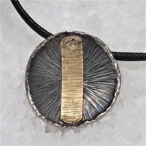 May include: A silver pendant with a gold bar in the center. The pendant has a textured surface and is attached to a black cord.