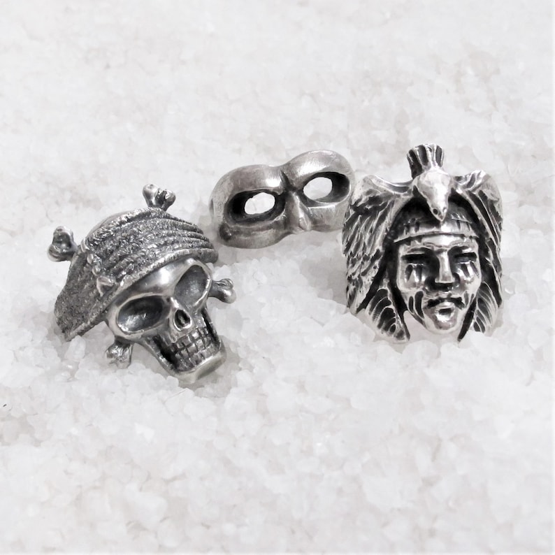 May include: Three silver-toned rings, one with a skull design, one with a Native American headdress design, and one with a mask design.