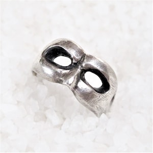 Op de afbeelding: Een zilveren ring in de vorm van een masker met twee ooggaten.