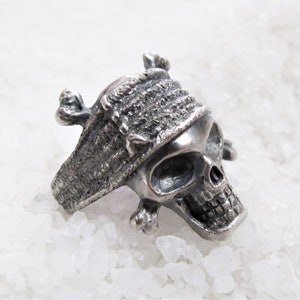 May include: A silver skull ring with a textured band and a skull wearing a bandana. The skull has a detailed face with a prominent nose and jawline.