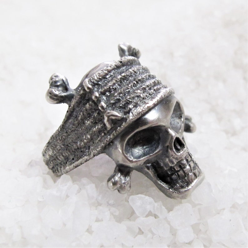 May include: A silver skull ring with a textured band and a skull wearing a bandana. The skull has a detailed face with a prominent nose and jawline.