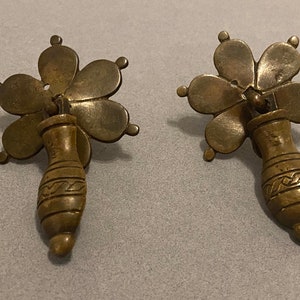 May include: Two antique brass drawer pulls. Each pull features a flower-shaped top with five petals and a decorative handle. The handles have a vase-like shape with etched patterns. The pulls are made of a brass-colored metal.