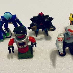 May include: A collection of six colorful robot action figures. The robots are in various poses and feature a range of colors, including red, blue, orange, black, and gray. These toys are likely from a children's toy collection.