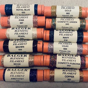 May include: A collection of twelve spools of Balger blending filament in various colors including royal blue, gold, pink, confetti green, pearl, navy, brown, lilac, beige, confetti fuchsia, amethyst, and copper, each labeled with its color and number.