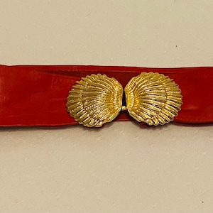 May include: A red leather belt with a gold-colored seashell clasp. The belt is wide and has a smooth texture. The clasp is made of two detailed seashell halves that connect in the center.