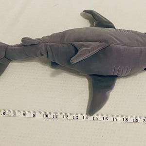 May include: A gray plush shark toy, approximately 24 inches long, lies on a white surface. The toy has fins, a tail, and small black eyes. A measuring tape is placed next to the shark.