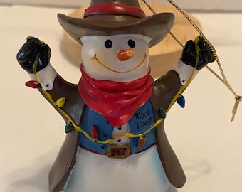 Pode incluir: Um enfeite de Natal de boneco de neve com chapéu e casaco de cowboy castanho, cachecol vermelho e colete azul. O colete tem a inscrição "Blood Donor". O boneco de neve é decorado com uma sequência de luzes coloridas.