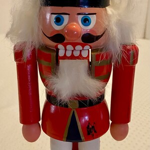 May include: A wooden nutcracker figurine in a red uniform with a black hat and boots. The figurine has a white beard and mustache, blue eyes, and a stern expression. It stands on a green base.