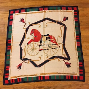 May include: Square silk scarf with a cream background featuring a red horse-drawn carriage design. The scarf has a navy blue, red, and green border with gold accents and tassels. The scarf is lying on a wooden surface.
