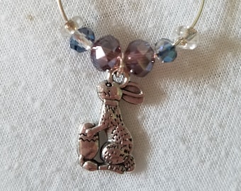 May include: A silver wine glass charm featuring a silver bunny holding an egg charm. The charm is adorned with purple and blue beads.
