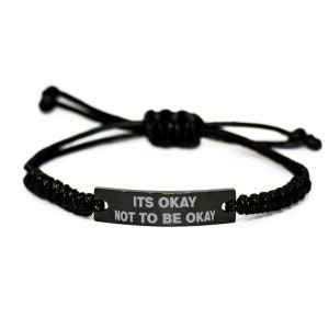 Puede incluir: Pulsera negra ajustable con una placa metálica rectangular. La placa tiene las palabras "IT'S OKAY NOT TO BE OKAY" en letras blancas. La pulsera está hecha de cordón negro tejido.