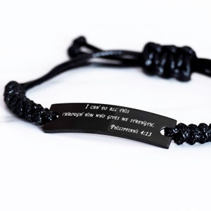Puede incluir: Pulsera trenzada negra con una etiqueta de metal negro que dice "Puedo hacerlo todo por medio de aquel que me fortalece. Filipenses 4:13"