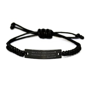 Pode incluir: Pulseira trançada preta com uma etiqueta de metal preto que diz "ABOVE ALL ELSE, GUARD YOUR HEART. FOR EVERYTHING YOU DO FLOWS FROM IT. PROVERBS 4:23".