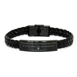 Puede incluir: Pulsera trenzada de cuero negro con una placa de metal negro grabada con el versículo bíblico "FOR THE SPIRIT GOD GAVE US DOES NOT MAKE US TIMID, BUT GIVES US POWER, LOVE AND SELF-DISCIPLINE. 2 TIMOTHY 1:7" y una pequeña cruz.