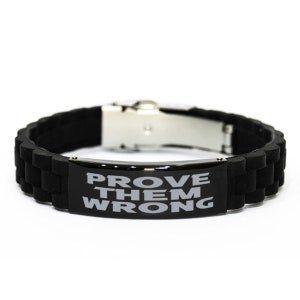 Puede incluir: Pulsera de silicona negra con cierre plateado y una placa de metal negro que dice "PROVE THEM WRONG" en letras grises.