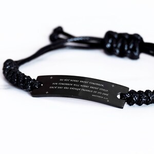 May include: Black bracelet with a woven cord design and a rectangular metal plate. The plate features the text "DO NOT WORRY ABOUT TOMORROW" and a Bible verse reference. The bracelet is on a white background.