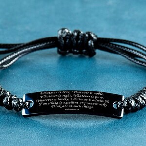 Pode incluir: Pulseira de cordão trançado preto com uma etiqueta de metal preto gravada com a citação "Whatever is true, Whatever is noble. Whatever is right, Whatever is pure, Whatever is lovely, Whatever is admirable If anything is excellent or praiseworthy Think about such things. Philippians 4:8"