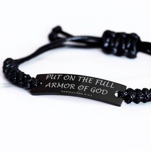 Puede incluir: Pulsera trenzada negra con una etiqueta de metal negro que dice "PUT ON THE FULL ARMOR OF GOD EPHESIANS 6:11"