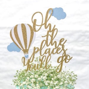 May include: A two-tiered cake with blue frosting and white sprinkles. The cake is decorated with a gold glitter hot air balloon and a topper that says "Oh the places you'll go".