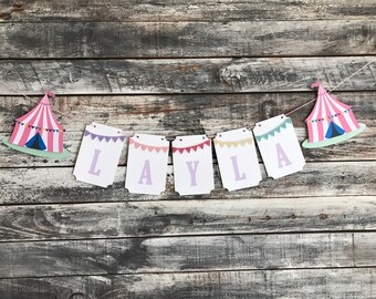 May include: A pink and white circus tent banner with the name "LAYLA" spelled out in purple letters. The banner is strung with a white string and has small colorful flags.