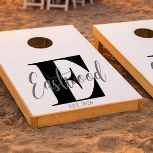 Op de afbeelding: Twee witte cornhole-borden met zijkanten van natuurlijk hout, elk voorzien van een grote zwarte letter E, het woord Eastwood in cursief schrift en de tekst EST. 2026. De borden staan op een zandstrand met een bruiloft bij zonsondergang op de achtergrond.