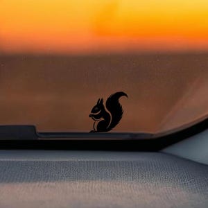 May include: A black squirrel decal is affixed to a car windshield. The squirrel is in a sitting position, holding a nut. The background features a sunset with orange and yellow hues.