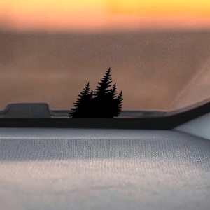 Op de afbeelding: Een zwart silhouet van dennenbomen op het dashboard van een auto, met een vage zonsondergang op de achtergrond. Het dashboard is grijs en de voorruit is helder. De lucht gaat over van geel naar oranje.