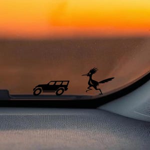 May include: Black vinyl decal of a vintage car and a roadrunner on a car windshield. The background is a sunset with orange and yellow hues. The dashboard is gray.