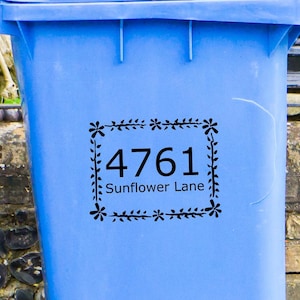 Op de afbeelding: Een blauwe recyclebak met een zwart-witte sticker waarop "4761 Sunflower Lane" staat in een decoratief bloemenframe.