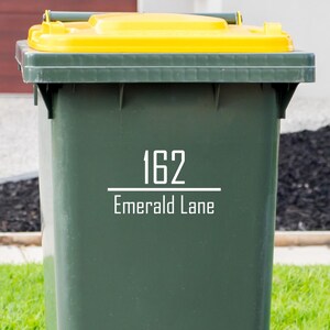Op de afbeelding: Een donkergroene afvalbak met een gele deksel. De bak heeft het adres "162 Emerald Lane" in witte tekst. De bak staat op een groen gazon.