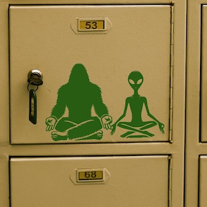 Könnte beinhalten: Nahaufnahme einer Reihe beiger Schließfächer mit grünen Aufklebern eines Bigfoot und eines Außerirdischen in meditativer Pose. Jedes Schließfach hat einen Schlüssel im Schloss. Der Bigfoot sitzt im Schneidersitz, und der Außerirdische befindet sich im Lotussitz.