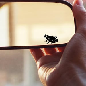 May include: A black frog decal is affixed to a car's rearview mirror. The frog is a simple silhouette, and the mirror is held by a hand. The background is blurred, suggesting the car's interior and exterior.