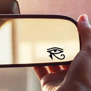 Peut inclure: Un autocollant noir représentant l'œil d'Horus est apposé sur le rétroviseur d'une voiture. Le miroir reflète une vue floue de l'intérieur et de l'extérieur de la voiture. Une main tient le miroir.