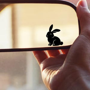 May include: A black rabbit decal is affixed to a car's rearview mirror. The decal is a silhouette of a rabbit in a sitting position. The mirror is held by a hand, and the background is blurred.