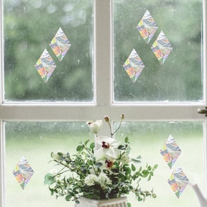 Könnte beinhalten: Ein Fenster mit bunten, rautenförmigen Aufklebern auf dem Glas. Das Fenster ist weiß gerahmt und hat eine weiße Fensterbank. Auf der Fensterbank stehen weiße Blumen in einer weißen Vase.