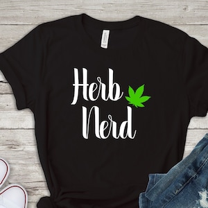Op de afbeelding: Zwart T-shirt met witte tekst die "Herb Nerd" zegt en een groene marihuana-bladgrafiek.