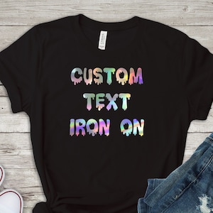Puede incluir: Camiseta negra con un diseño de plancha holográfico que dice "CUSTOM TEXT IRON ON".