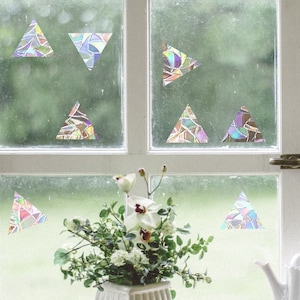 Puede incluir: Una ventana con marco blanco y una vista verde al exterior. Hay varias pegatinas triangulares iridiscentes en la ventana. Un ramo de flores blancas está en un jarrón blanco en el alféizar de la ventana.