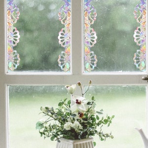 Op de afbeelding: Een wit raamkozijn is voorzien van verticale stroken iriserende raamfolie in glas-in-loodstijl met een herhalend bloemmotief. De folie vertoont een kleurenspectrum met paars, oranje en groen dat het licht opvangt.