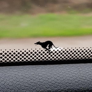 Pode incluir: Silhueta preta de um galgo a correr, fixada no painel do carro. O cão está posicionado acima de uma faixa de ventilação perfurada, com um fundo desfocado verde e castanho visível.