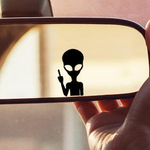 Puede incluir: Silueta negra de un extraterrestre haciendo una peineta, adherida al espejo retrovisor de un coche. La imagen está sobre un fondo borroso, con una mano sujetando el espejo.