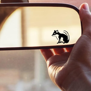 May include: A black and white sticker of a small animal, possibly a rodent, is affixed to a car's rearview mirror. The sticker is a simple silhouette design. The mirror is held by a person's hand, with the car's windshield visible in the background.