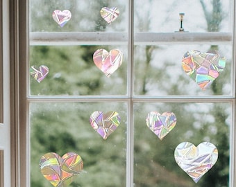 Puede incluir: Una ventana con ocho pegatinas en forma de corazón iridiscentes. Las pegatinas están hechas de un material transparente con un acabado similar al arcoíris. Los corazones son de diferentes tamaños y están dispuestos en un patrón aleatorio en la ventana.