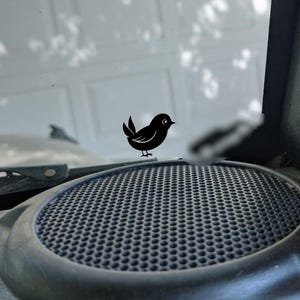 May include: A black, stylized bird sticker perched on a black speaker grill. The speaker is set into a dark gray dashboard. The background shows a blurred view of a window and a garage door.