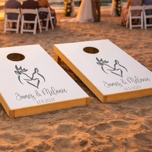Könnte beinhalten: Zwei weiße Cornhole-Boards mit Holzrahmen, die jeweils eine schwarze Grafik von zwei Hirschköpfen in Herzform, die Namen James & Melanie sowie das Datum 22.5.2027 in schwarzer Schrift auf der Oberfläche der Boards zeigen.