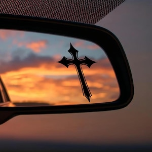 May include: A black cross-shaped decal is attached to a car's rearview mirror. The cross is set against a vibrant sunset sky with orange, yellow, and pink hues. The image is framed by the car's interior.