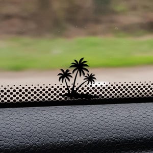 以下が含まれることがあります： 3本のヤシの木が描かれた黒いヤシの木のステッカー。ステッカーは車の窓に貼られており、その下に穴の開いたストリップがあります。背景にはぼやけた緑の風景が写っています。
